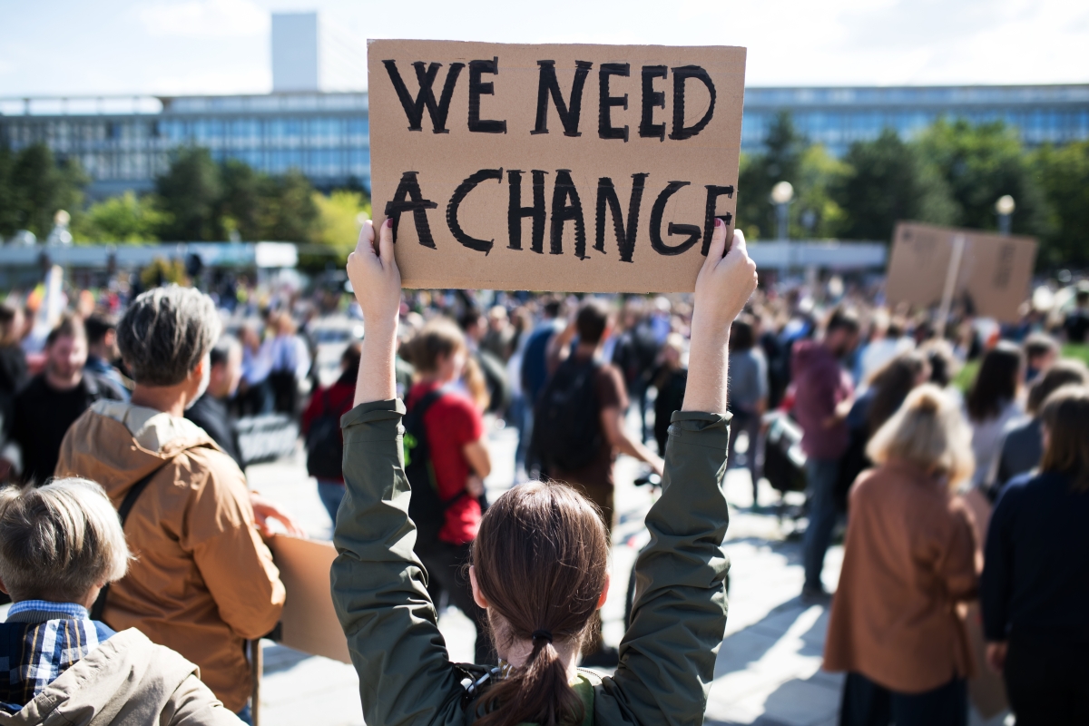 Blick auf eine Protestveranstaltung, viele Menschen von hinten, mittig steht eine Frau und hält ein Plakat mit der Aufschrift "We need a change" hoch